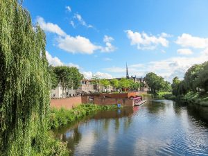 a river as seen from a bridge with a weeping willow on the left and church spire at the background , in the middle a cafe with lots of tables at the river bank on a platform, De Dommel River, Den Bosch