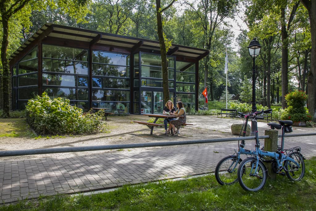 a building made of glass in a forest with 2 women sitting at a table outside and 2 bikes parked in front; the Tourist information point in Diever, Drenthe