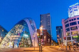 modern glass building with a rounded form in front of skyrises in the night lit up in colors; Things to do Eindhoven