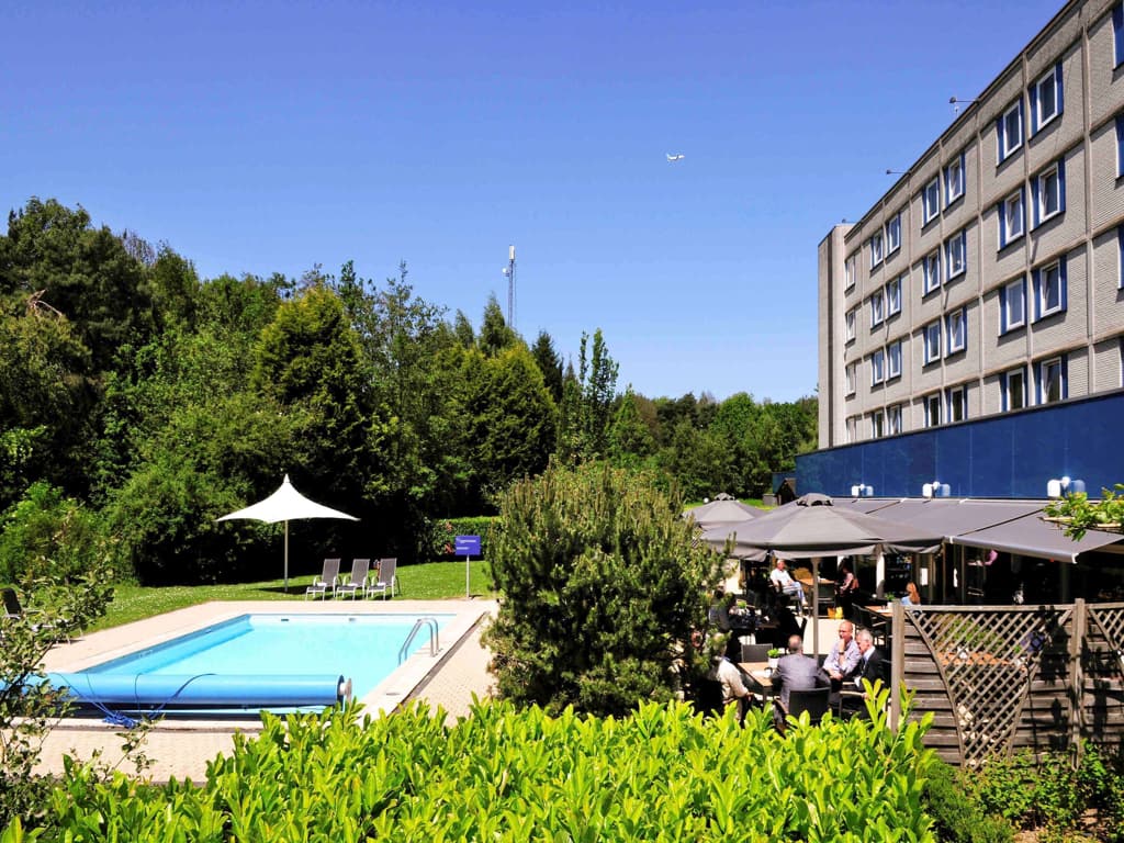 A hotel with a swimming pool surrounded by lush trees in the background, Novotel in Eindhoven