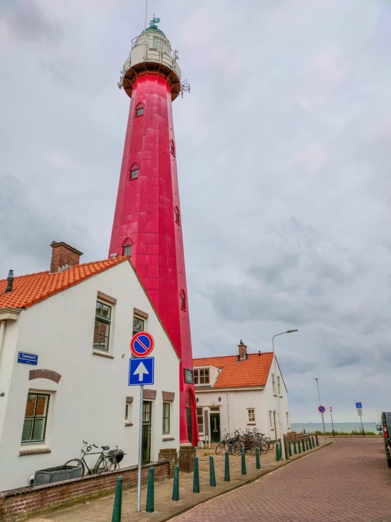 a red lighthouse with a white top between two white buildings, at the background - a bit of a sea; on a gloomy day with dark rain clouds; The Lighthouse of Scheveningen