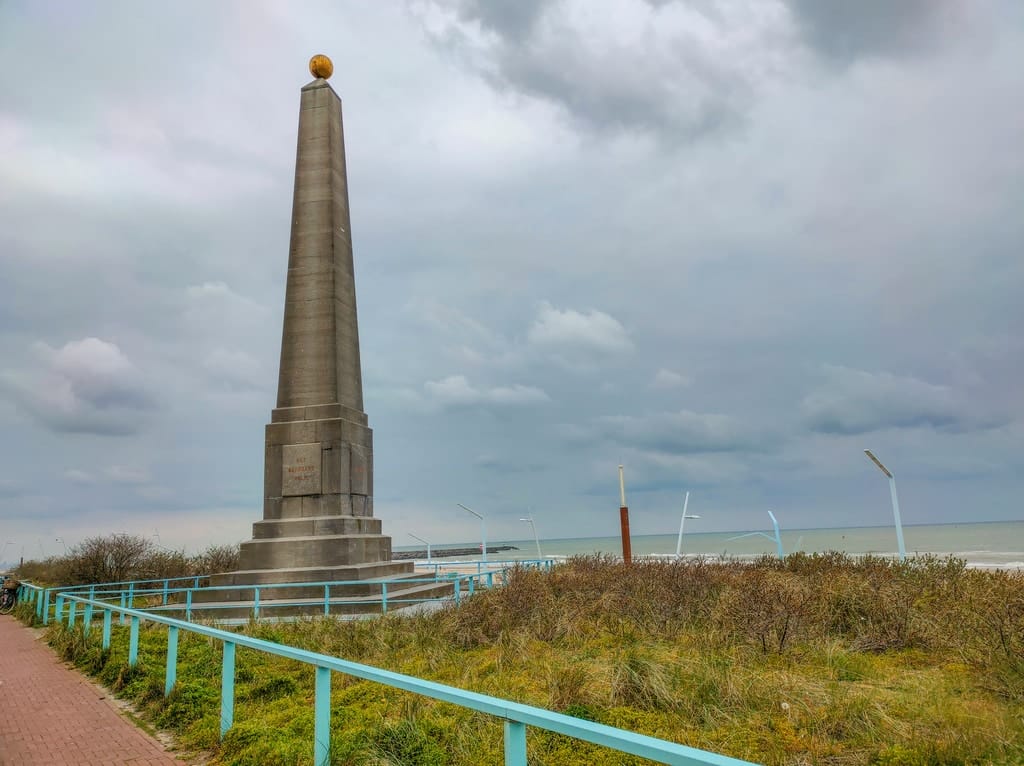an obelisk with a golden globe on the top, at the background - sea; on a gloomy day with dark clouds; The Memorial Needle in Scheveningen