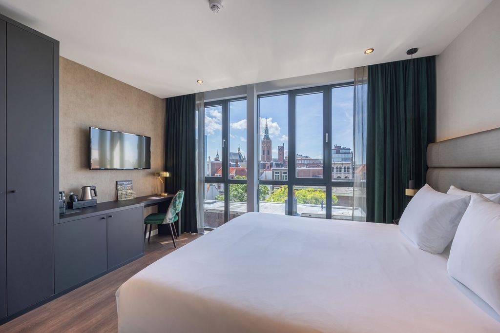 A modern hotel room with a large bed, a desk, and a window with a view of a cityscape. The bed has white linens and two pillows. The desk has a chair with a green floral pattern, a lamp, a coffee maker, and a small tray with cups. Above the desk, there is a flat-screen TV mounted on the wall. A room at Boutique Hotel Corona in The Hague