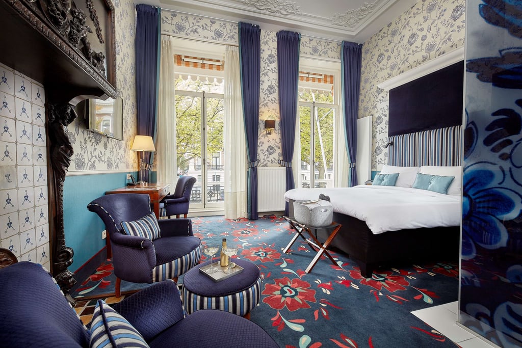 A well-decorated hotel room or a luxurious bedroom. The room features a large bed with a dark blue headboard and several light blue pillows. There are two large windows with sheer white curtains and dark blue drapes, allowing natural light to enter the room. The walls are adorned with floral wallpaper, and the ceiling has intricate moldings. A room at the Carlton Ambassador Hotel in The Hague
