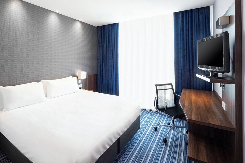 The image shows a modern, well-lit hotel room with a minimalist design. The room features a large bed with white linens and two pillows, positioned against a textured gray wall. There is a small bedside table with a lamp on the left side of the bed. On the right side of the room, there is a wooden desk with a flat-screen television mounted on the wall above it. A black office chair with a mesh back is placed in front of the desk. The floor is covered with a blue and white striped carpet, and the window is dressed with white sheer curtains and dark blue blackout curtains. The room appears clean and organized, with a focus on functionality and comfort. A room at the Holiday Inn Express Hotel in The Hague