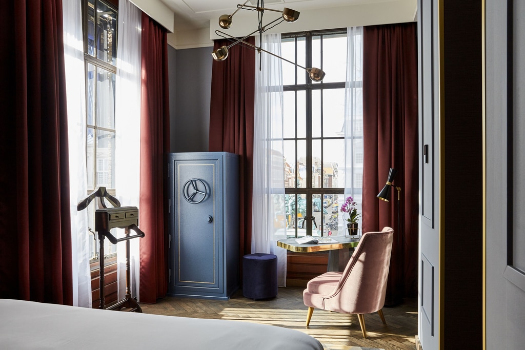 The image shows a well-decorated room with a large window that allows natural light to enter. The room features dark red curtains, a blue safe with a circular handle, a small round table with a marble top, a pink upholstered chair, a floor lamp, and a valet stand. There is also a small purple ottoman next to the safe and a potted orchid on the table. A room at the Hotel Indigo in The Hague