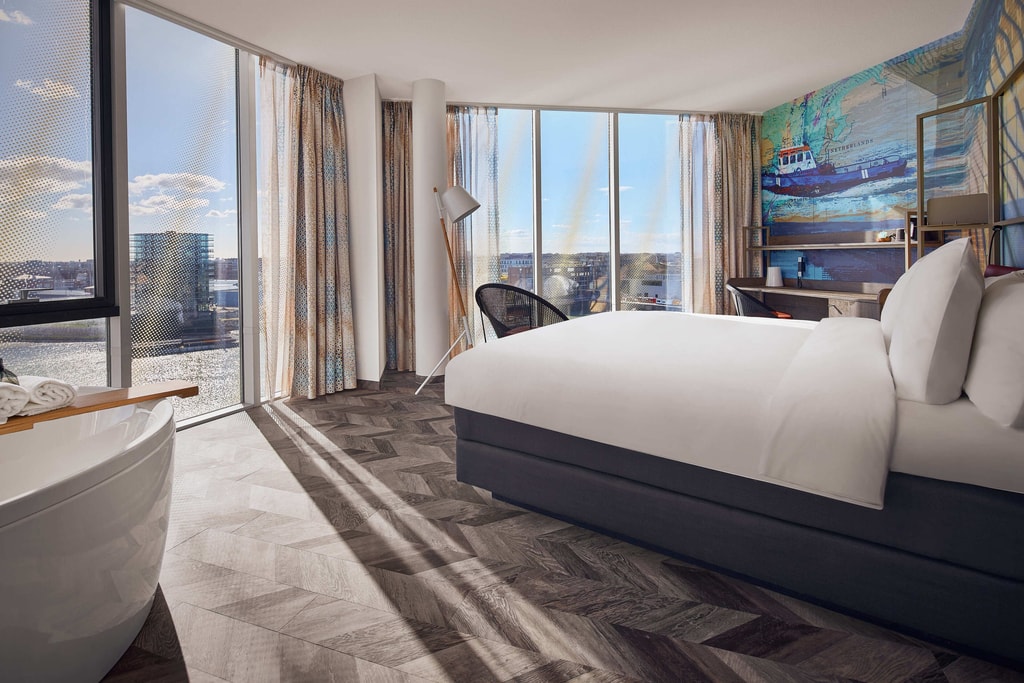 A modern, well-lit hotel room with large floor-to-ceiling windows that offer a view of a waterfront and cityscape. The room features a large bed with white linens, a freestanding bathtub with a wooden tray holding towels, and a stylish floor lamp. The walls are decorated with a colorful mural depicting a ship and a map, adding a nautical theme to the room. There is also a desk with a chair and some items on it, such as a lamp and a cup. The floor has a herringbone pattern, and the curtains are light-colored with a subtle pattern. The overall ambiance of the room is bright and inviting, with natural light streaming in through the windows. A room with a view at the Inntel Hotels Den Haag Marina Beach