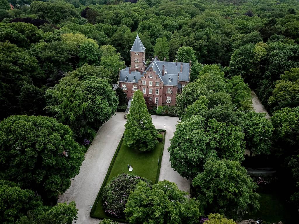 The image shows an aerial view of a large, historic-looking building surrounded by dense, lush green forest. The building appears to be a castle or a manor house with a tall, pointed tower and multiple chimneys. The structure is made of red brick with a grey slate roof. In front of the building, there is a well-maintained garden with a large, triangular-shaped lawn and a central pathway leading up to the entrance. The garden is bordered by neatly trimmed hedges and trees. There are also gravel paths on either side of the garden, leading through the forested area. The image captures the grandeur and isolation of the building, emphasizing its integration with the natural surroundings. Kasteel de Wittenburg in The Hague