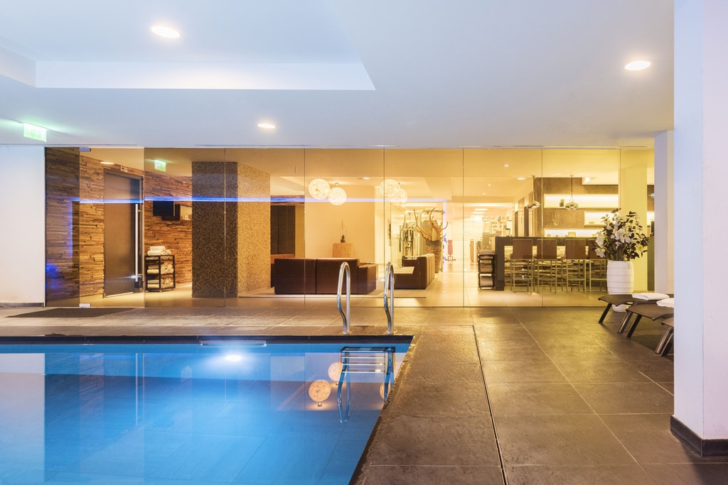 An indoor swimming pool area with a modern and luxurious design. The pool is in the foreground, with clear blue water and a metal ladder for entry. Surrounding the pool is a tiled floor with a dark, sleek finish. To the right of the pool, there are two lounge chairs with white towels placed on them, and a large potted plant with white flowers. The swimming pool at the Leonardo Royal Hotel Den Haag hotel