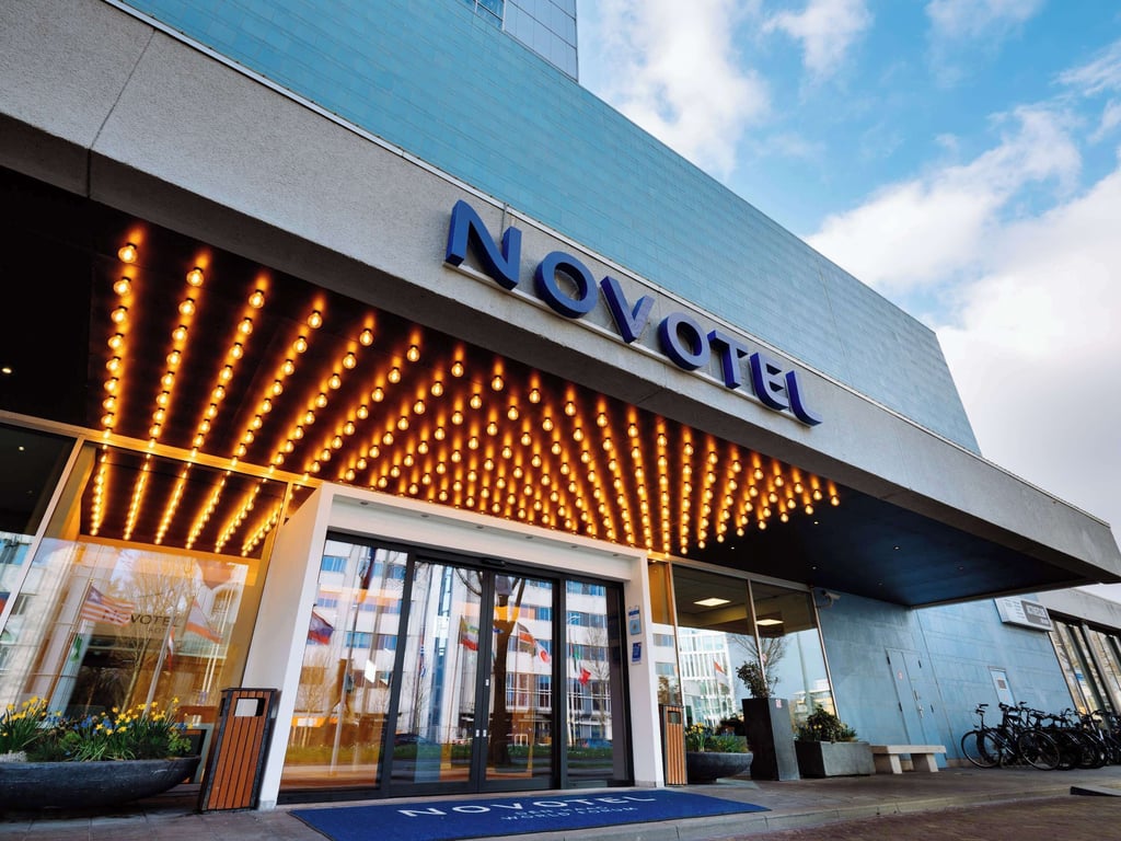 The exterior entrance of a Novotel hotel. The building has a modern design with a large sign displaying the hotel's name "Novotel" in bold, blue letters. Above the entrance, there is a canopy adorned with numerous small, round lights that are illuminated, creating a warm and inviting atmosphere. The entrance features large glass doors. Novotel Den Haag World Forum