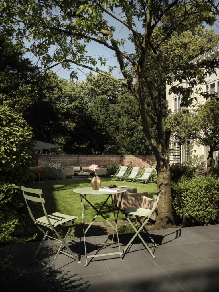 A serene and well-maintained backyard garden. In the foreground, there is a small round table with two matching chairs, all in a light green color. On the table, there is a vase with a single pink flower, a book, and a pair of sunglasses. The table and chairs are placed on a tiled patio area. The inner yard of Park Centraal Den Haag Hotel