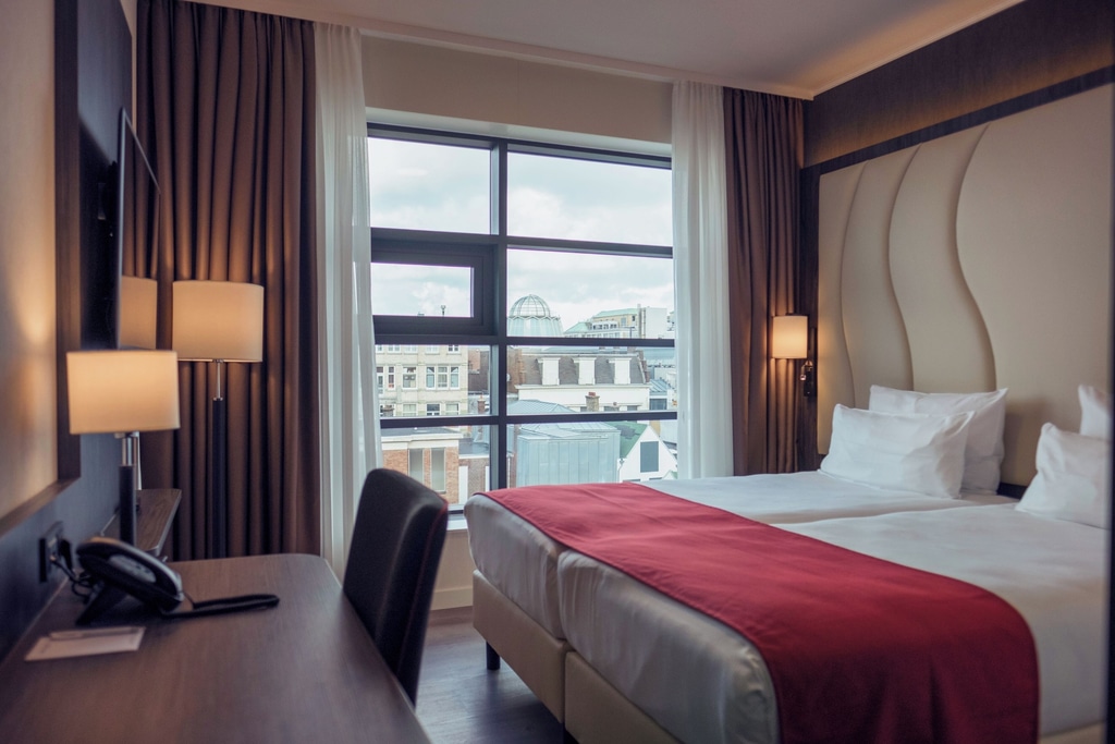 A well-furnished hotel room with a modern and clean design. The room features two single beds pushed together, each with white bedding and pillows, and a red runner across the foot of the beds. The headboard has a stylish, wavy design. There are two bedside lamps on either side of the bed, providing warm lighting. A room at the Best Western Plus Plaza Den Haag City Center