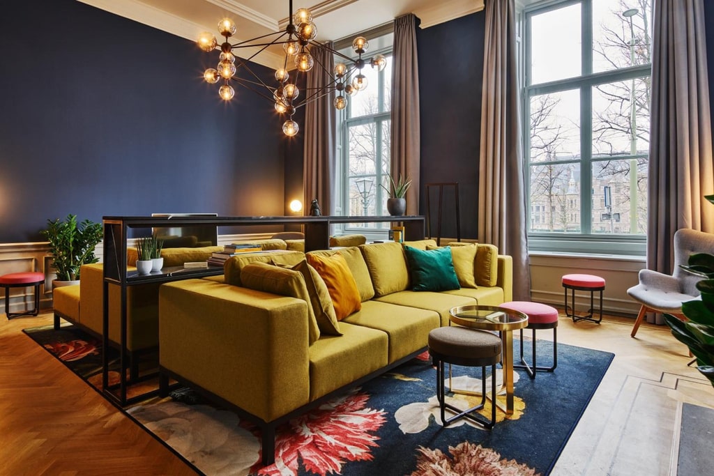 The image shows a modern, stylish living room with a sophisticated design. The room features a large mustard yellow sectional sofa adorned with various colored cushions, including green, yellow, and teal. The sofa is placed on a dark floral-patterned rug that adds a touch of elegance to the space. In front of the sofa, there are two small round tables with metallic frames and glass tops, accompanied by a small ottoman. A living room at the Staybridge Suites in The Hague