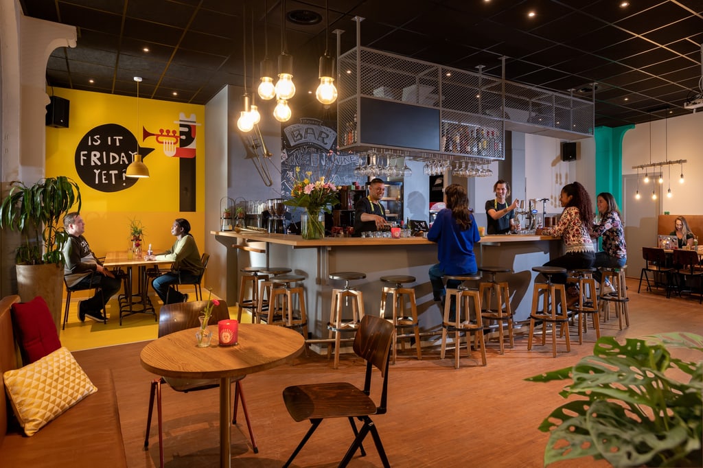 The image shows a modern, cozy bar or café. The space is well-lit with a combination of natural and artificial lighting. There are several people seated at the bar and at tables, engaging in conversation and enjoying drinks. The bar area is central in the image, with a bartender serving drinks. The bar has a variety of glasses hanging overhead and bottles displayed on shelves. There are several stools around the bar, and a few tables with chairs scattered around the room. A room at the Stayokay Hostel in The Hague