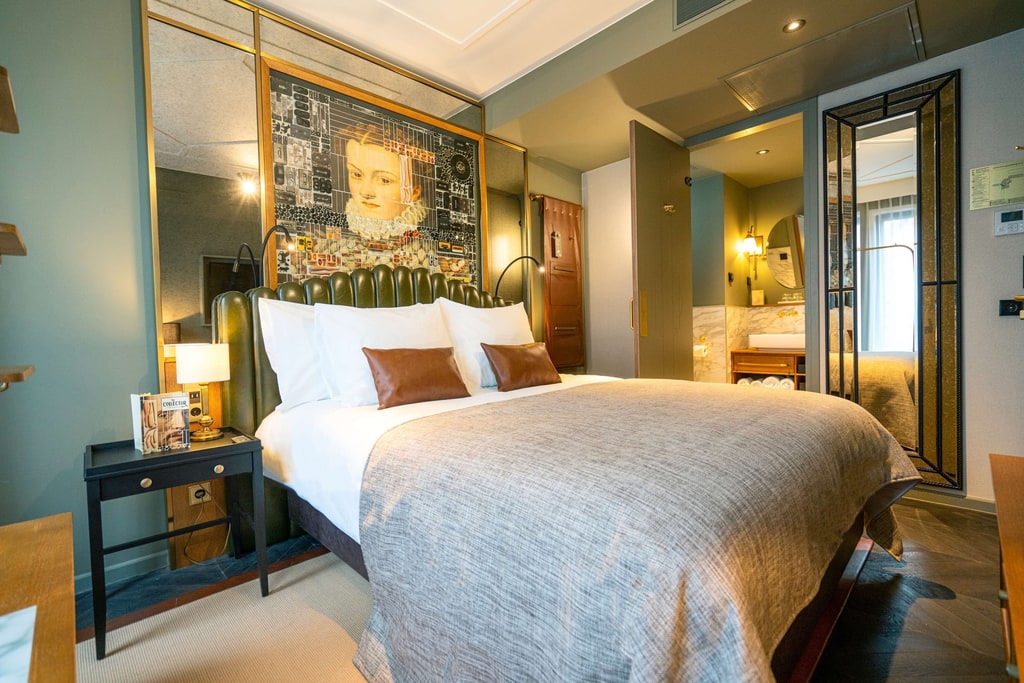 A well-decorated hotel room with a modern and stylish design. The room features a large bed with white linens, two brown accent pillows, and a grey bedspread. The headboard is upholstered in green leather. On the left side of the bed, there is a black nightstand with a lamp and a book titled "Ceviche" on it. The wall behind the bed has a large decorative panel with a complex, circuit-like design. A room at The Collector Hotel in The Hague