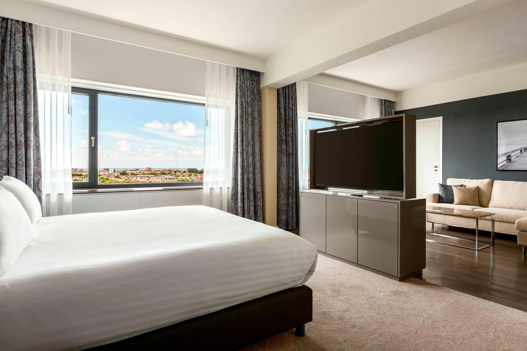 A modern, well-lit hotel room with a large window offering a scenic view of a cityscape and a blue sky with scattered clouds. The room features a neatly made bed with white linens on the left side, a large flat-screen TV on a stand in the center, and a sitting area with a beige sofa and a glass coffee table on the right side. A room at The Hague Marriott Hotel