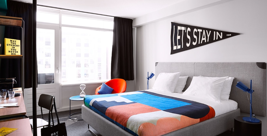 The image shows a modern, well-decorated bedroom with a large window that allows natural light to enter the room. The bed has a grey upholstered headboard and is made with white pillows and a colorful geometric-patterned blanket. On either side of the bed, there are blue reading lamps on small round tables. Above the bed, there is a black pennant with white text that reads "LET'S STAY IN". A hotel room in The Social Hub The Hague