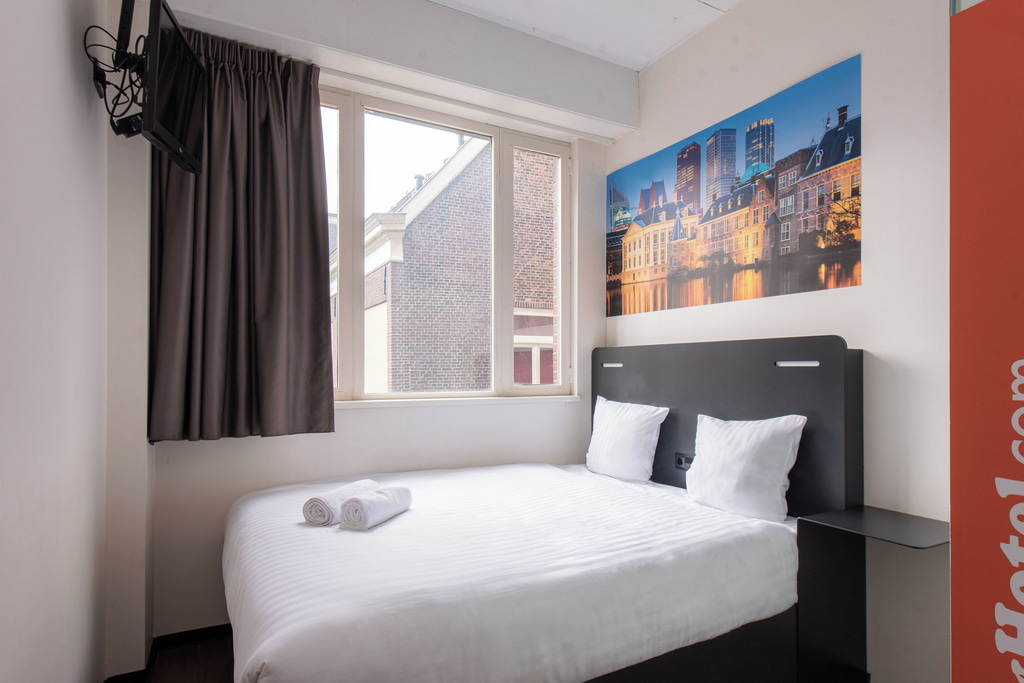 A small, neatly arranged hotel room. The room features a bed with white linens and two pillows, along with two rolled white towels placed on the bed. There is a window with dark curtains partially drawn, revealing a view of a brick building outside. A wall-mounted television is visible in the upper left corner. A room at the easyHotel in The Hague