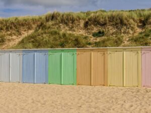 A row of charming beach huts arranged along a sandy shore, each painted in soft pastel shades like white, blue, green, orange, yellow, and pink. Behind the huts, grassy dunes rise gently toward a partly cloudy sky. Beach towns in the Netherlands
