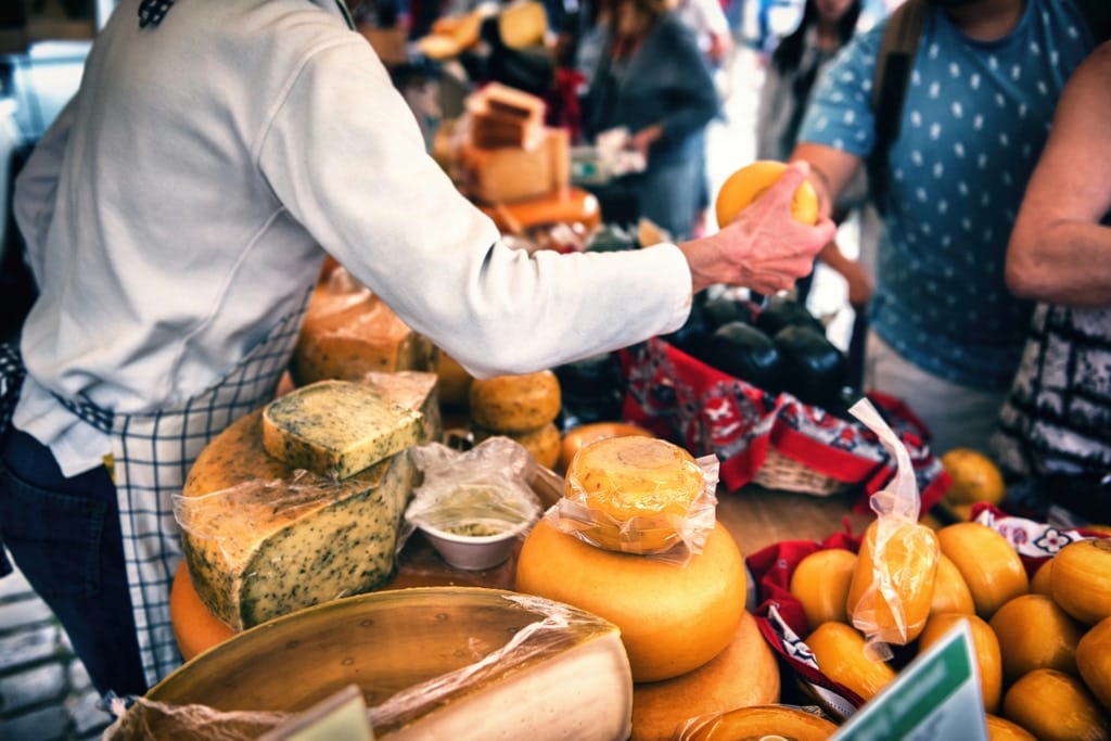 A vendor hands a round of cheese to a customer at an outdoor market stall with various blocks and wheels of cheese on display. Other shoppers and market goods are visible in the background. Shopping during a food tour in Amsterdam