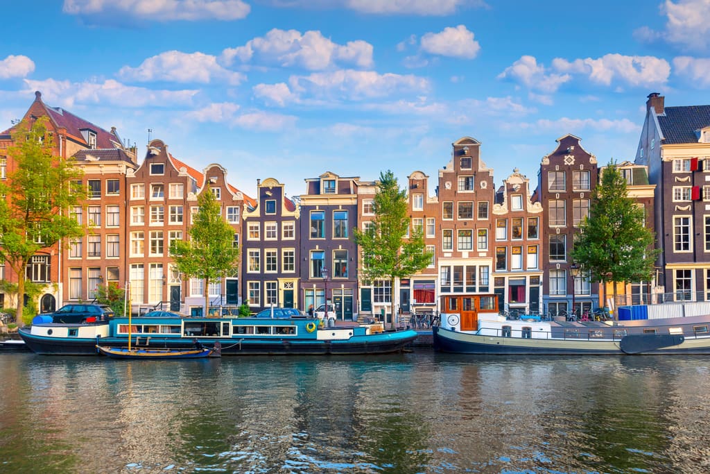 Colorful, narrow canal houses line the waterfront in Amsterdam, Netherlands. Several houseboats are moored along the edge of the calm canal, and trees are spaced between the buildings under a blue sky with clouds.
