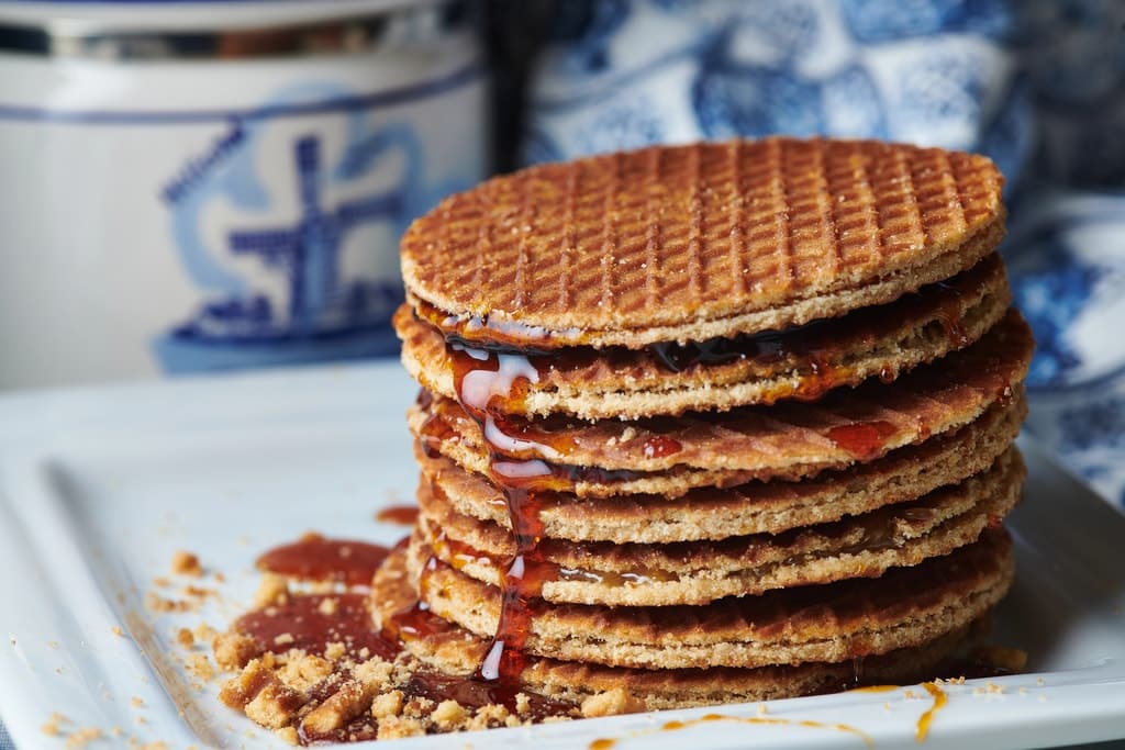 A stack of stroopwafels with syrup dripping between the layers sits on a white plate, with some crumbs scattered around and a blue and white decorative jar in the background. Dutch stroopwafels