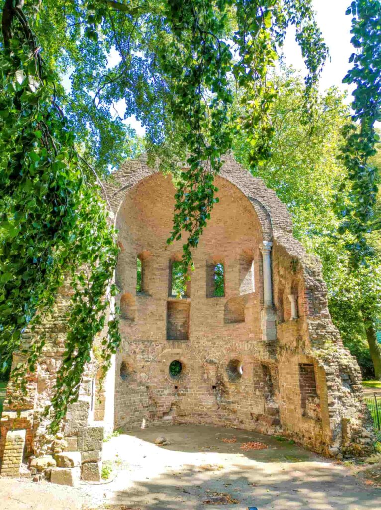 Ancient brick ruin with arched ceiling and several small window openings, surrounded by lush green trees on a sunny day. Barbarossa ruins in Valkhof Park, Nijmegen