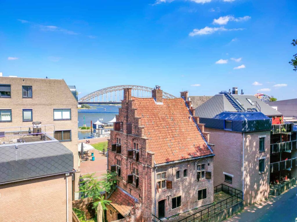 A view of a historic brick building with a red-tiled roof, modern buildings nearby, and a large arched bridge crossing a river in the background under a blue sky. Besiendershuis in Nijmegen