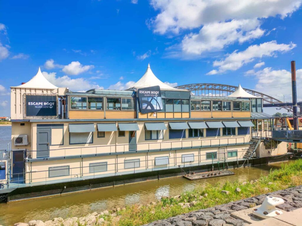 A large houseboat labelled “Escape Boot” is moored by a river under a partly cloudy blue sky, with a bridge and grassy bank visible nearby.