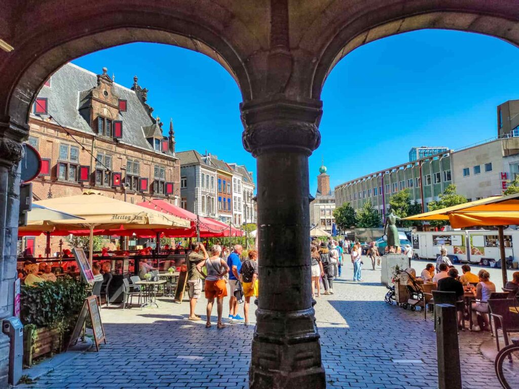 View of a lively outdoor square through two stone arches, with people walking, colourful umbrellas, historic buildings, and a bright blue sky. Grote Markt through Kerkboog, Nijmegen
