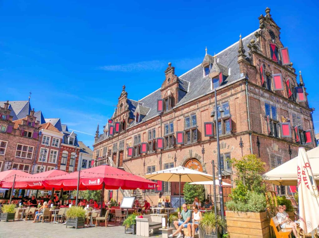 People sit at outdoor tables under red umbrellas in front of a historic brick building with decorative windows and a steep roof on a sunny day. The sky is clear and blue. Grote Markt with De Waagh in Nijmegen