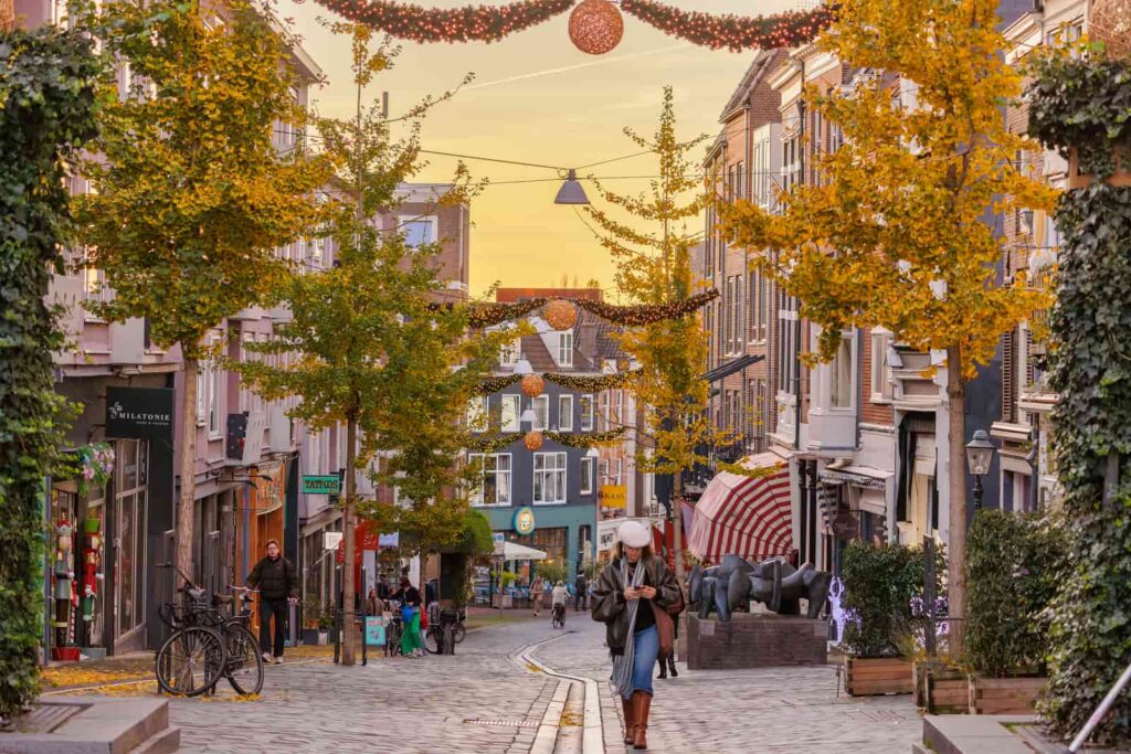 A person walks down a cobbled street lined with shops, trees with autumn leaves, and festive hanging decorations at sunset. Other people can be seen in the distance. Lange Hezelstraat in Nijmegen