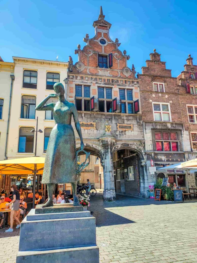 A bronze statue of a woman stands in a cobbled square near outdoor café seating, in front of ornate, historic brick buildings under a bright blue sky. Mariken van Nieumeghen Statue in Nijmegen
