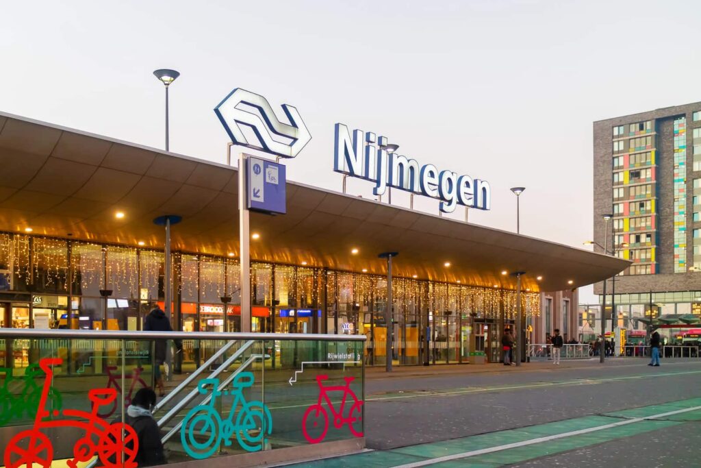 Nijmegen railway station entrance with large sign, festive lights hanging from the roof, bicycle-themed glass panels, and a modern building in the background at dusk. Nijmegen Central Train Station
