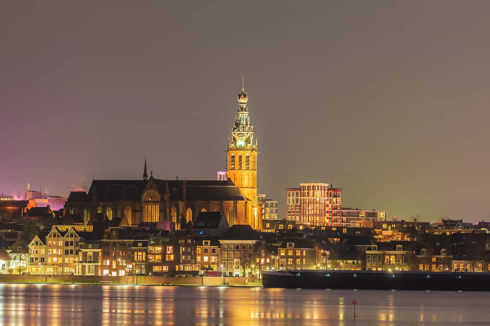 Night-time cityscape with illuminated historic buildings and a tall church tower reflecting on calm water, under a clear sky. Nijmegen, the Netherlands, at Night
