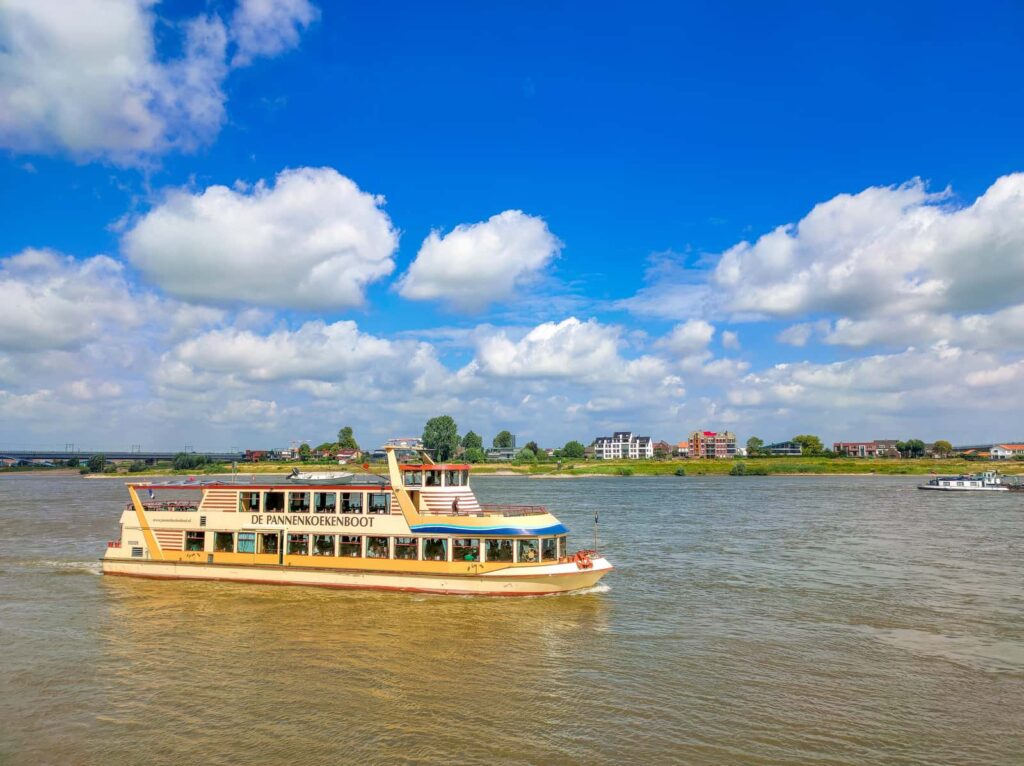 A yellow and white tour boat labelled “DE PANNEKOEKENBOOT” sails on a wide river under a partly cloudy sky, with buildings and trees visible on the distant shore.