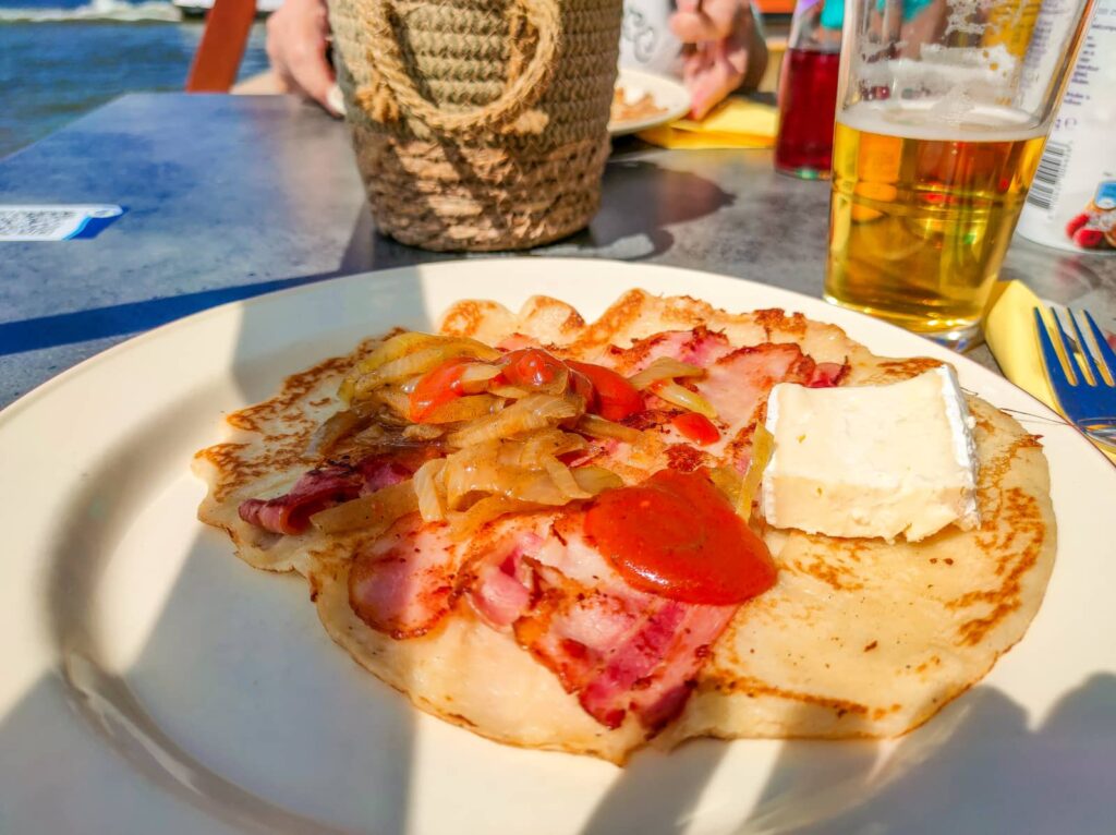 A savoury pancake topped with bacon, onions, tomatoes, and a slice of brie cheese, served on a plate outdoors with a glass of beer and a woven basket in the background.