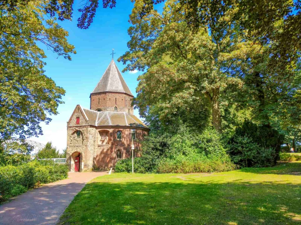 A small, historic brick church with a steep, pointed roof stands surrounded by green trees and grass under a bright blue sky on a sunny day. St Nicholas Chapel at Valkhof Park in Nijmegen