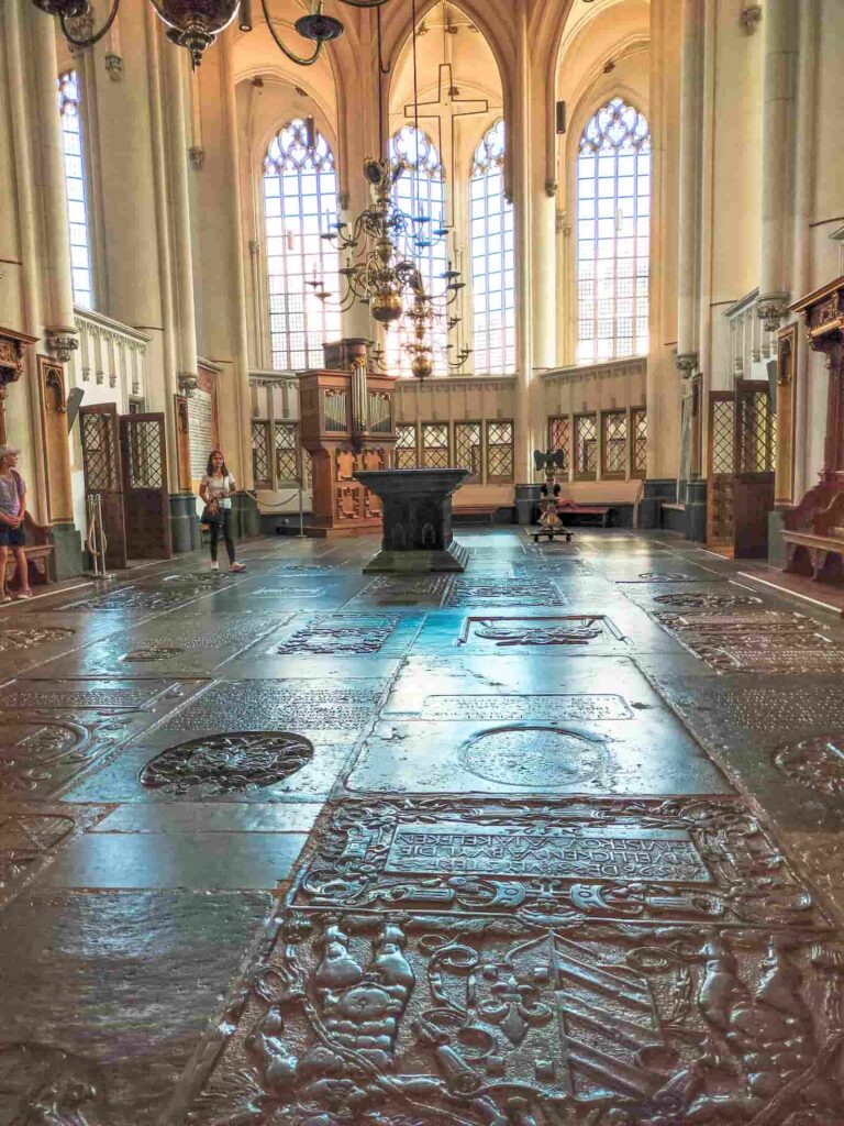 A historic church interior with ornate stone slabs on the floor, tall arched windows, chandeliers, and a few people sitting or standing near the walls. Inside Stevenskerk in Nijmegen
