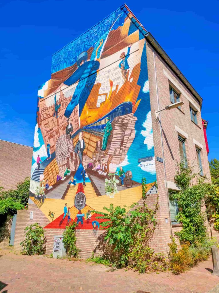 A colourful mural on a brick building depicts people, books, trains, and cityscapes, blending history and imagination with vibrant blues, oranges, and greens under a clear blue sky. Street art in Nijmegen