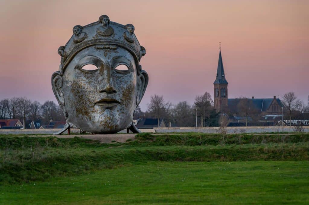 A large metallic face sculpture stands in a grassy field at sunset, with a distant village and church spire visible under a pink sky. The Face of Nijmegen sculpture
