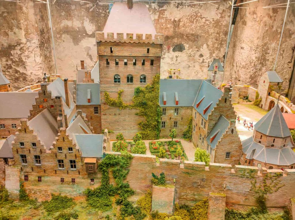 A detailed miniature model of a historic castle with stone walls, towers, and ivy climbing up the central building, set against a textured backdrop. Valkhof Castle model in St Nicholas Chapel, Nijmegen
