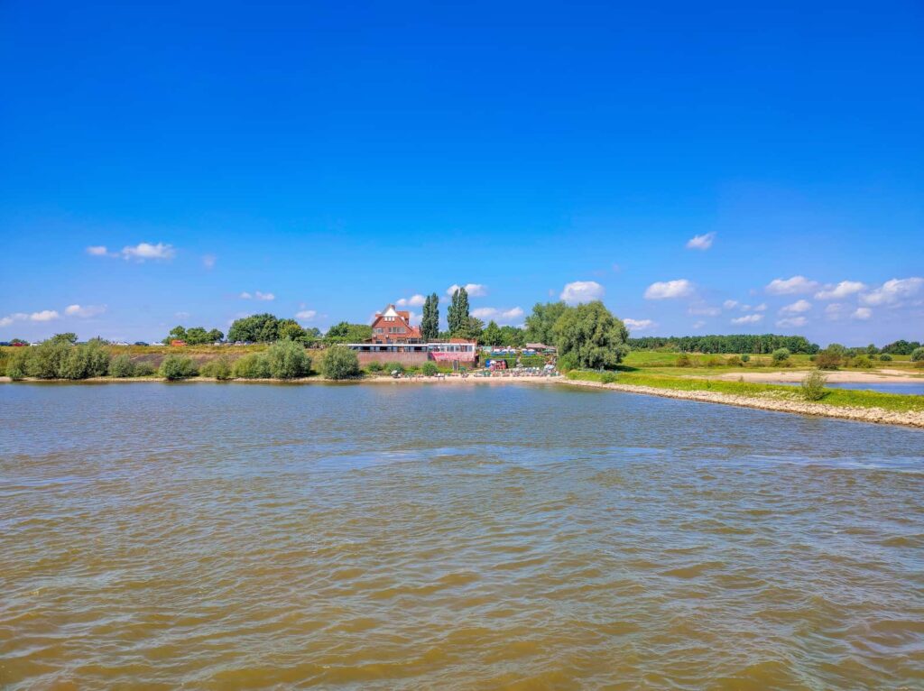 A wide river with a rocky shore, trees, and a red-brick building in the distance under a clear blue sky with scattered clouds. Veur-Lent island Nijmegen