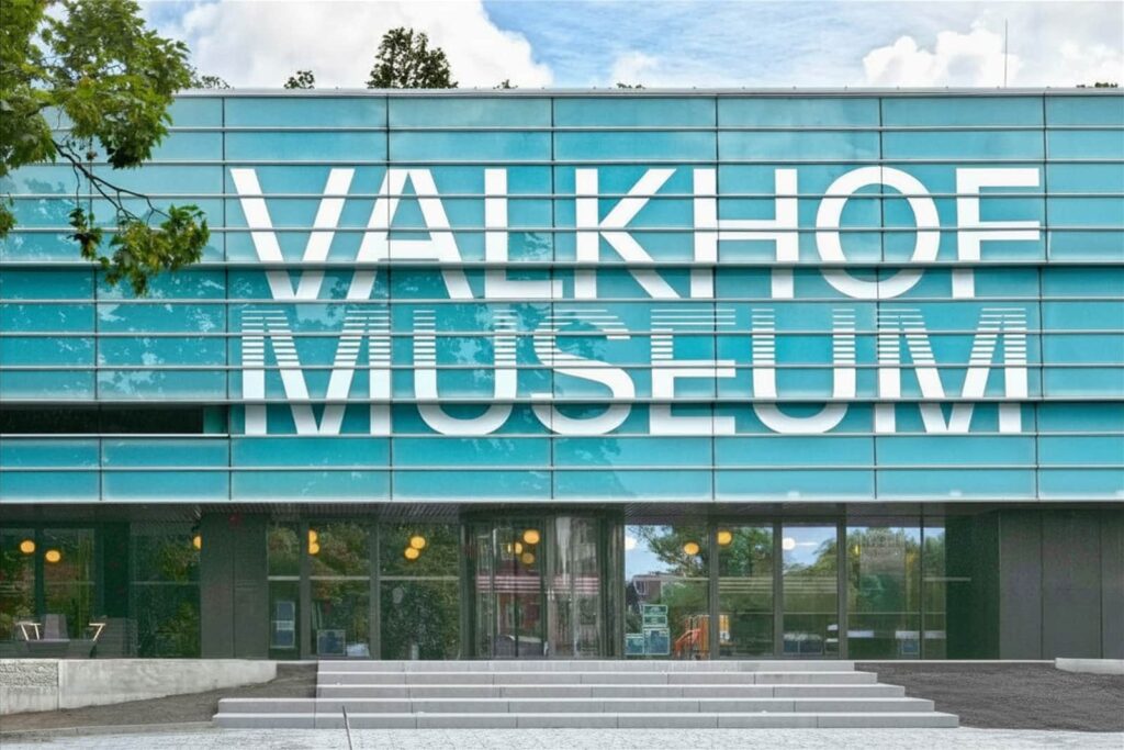 Modern glass building with large white text reading “Valkhof Museum” across the facade. Trees reflect in the windows, and the entrance is visible at the centre of the building.