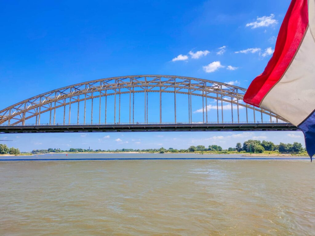 A large arched steel bridge spans a wide river under a blue sky, with a red, white, and blue flag partially visible on the right side of the image. Waalbrug from the Pancake Boat in Nijmegen