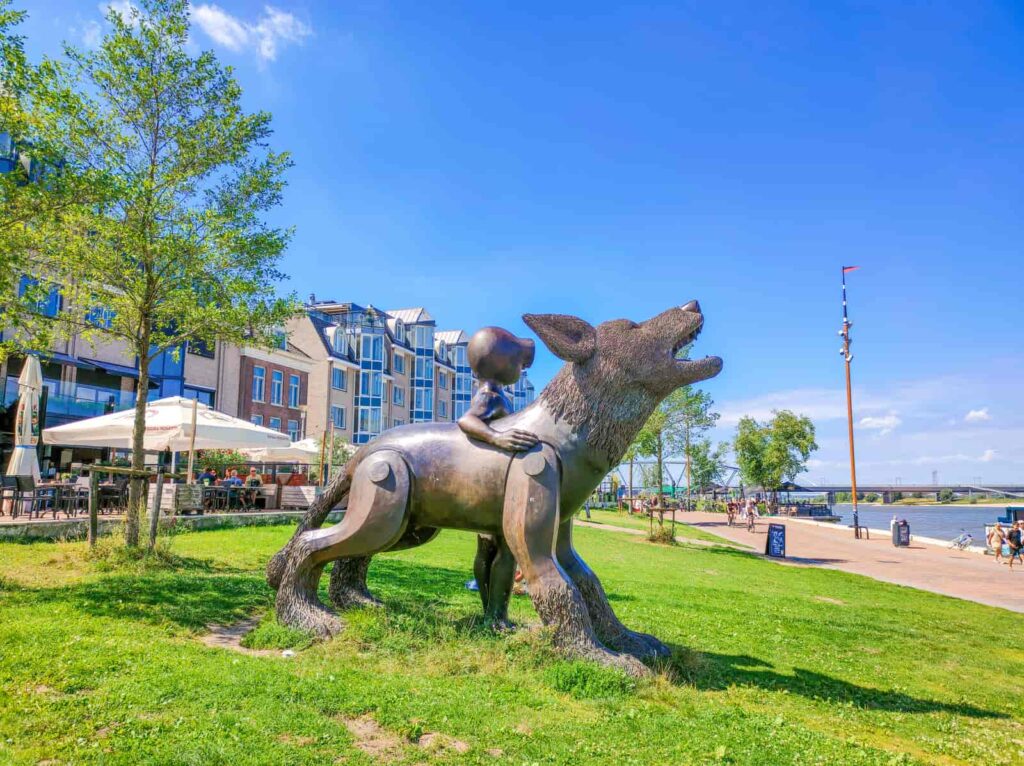Large bronze statue of a child next to a howling wolf stands on green grass near a riverside walkway, with people, flats, and trees visible under a clear blue sky. Waalkade in Nijmegen